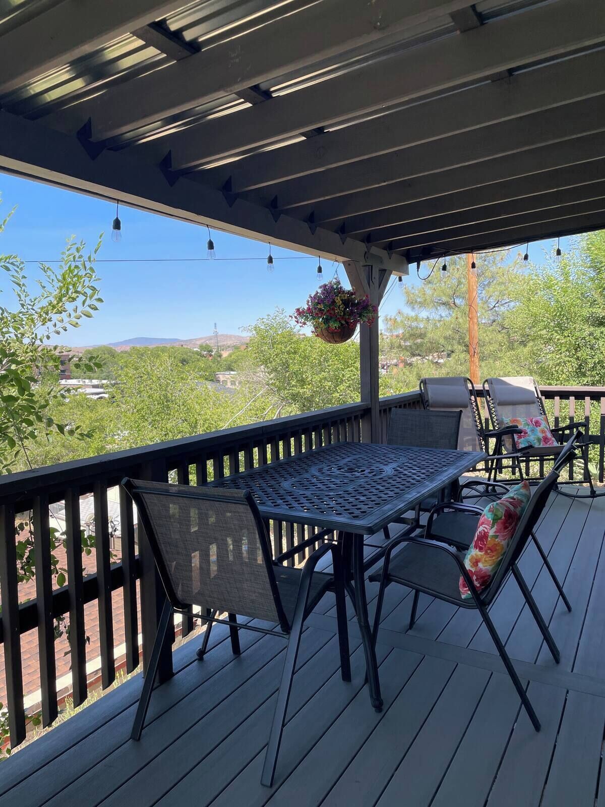 Une terrasse extérieure couverte, avec une table et des chaises, une jardinière suspendue et une vue sur un paysage verdoyant.
