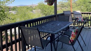 Un patio techado al aire libre con una mesa y sillas, una cesta colgante de flores y vistas a un paisaje verde.