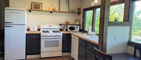 A compact kitchen with a refrigerator, stove, microwave, and a sink. There is a window providing natural light and a view of greenery outside.