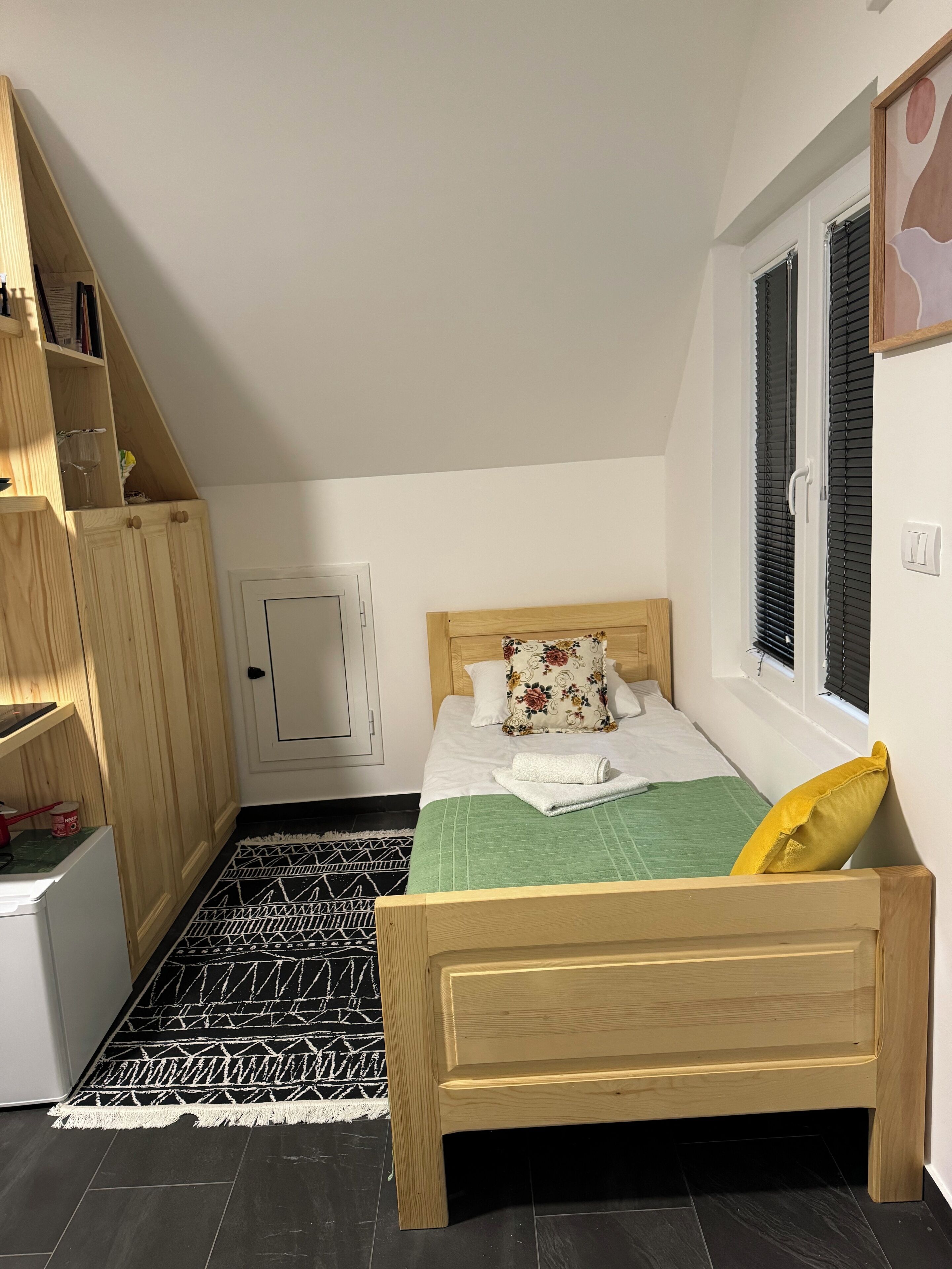 A bedroom with a wooden bed, a green bedspread, a yellow pillow, a wooden wardrobe, a small white fridge, a patterned rug, and a window with blinds.