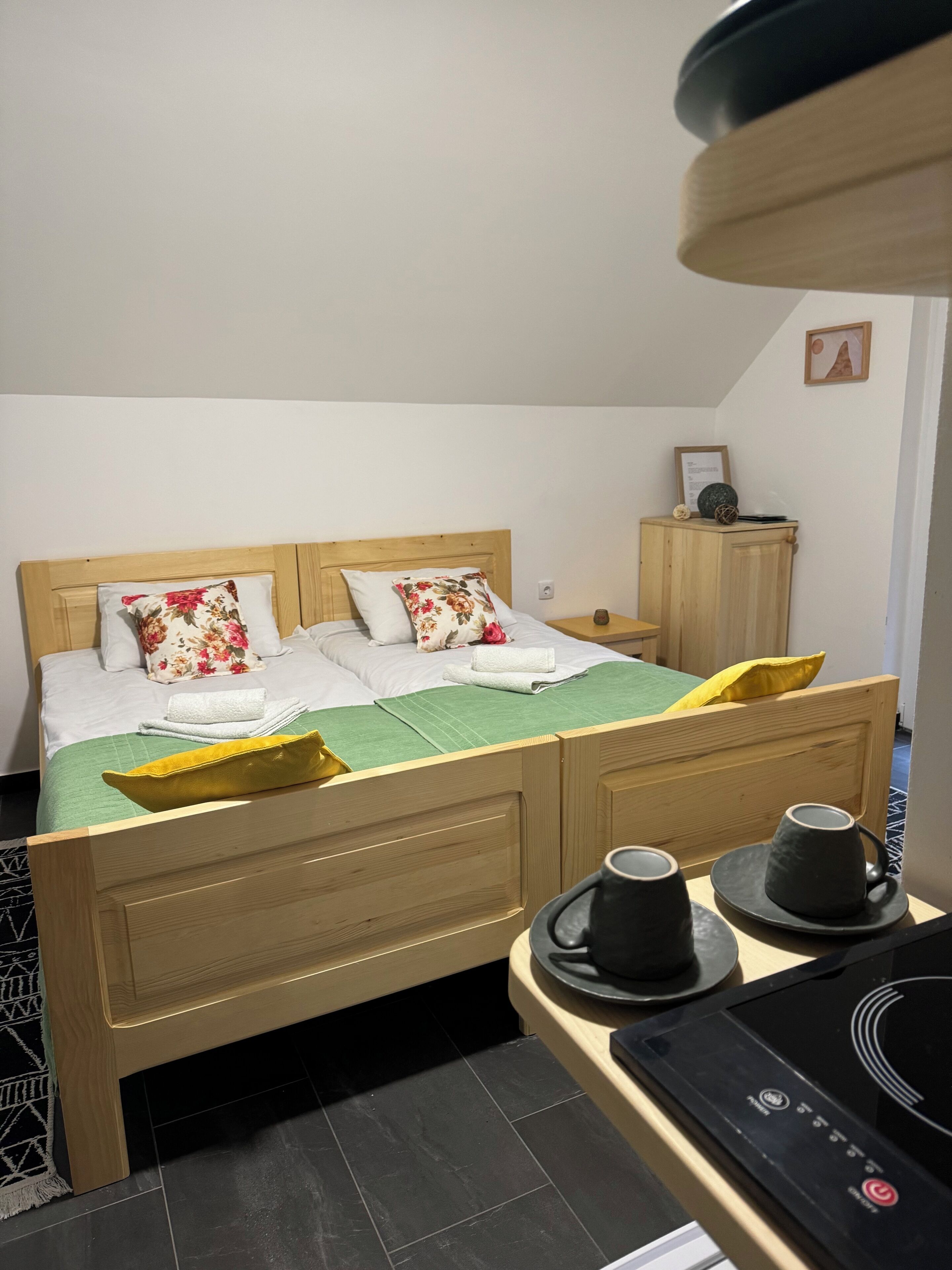 A neatly arranged hotel room with a wooden bed, green bedspread, floral pillows, a small wooden nightstand, and a coffee station with two cups.