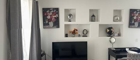 A living room with a flat-screen TV, a black entertainment unit, a dining area with a patterned chair, and decorative wall shelves.