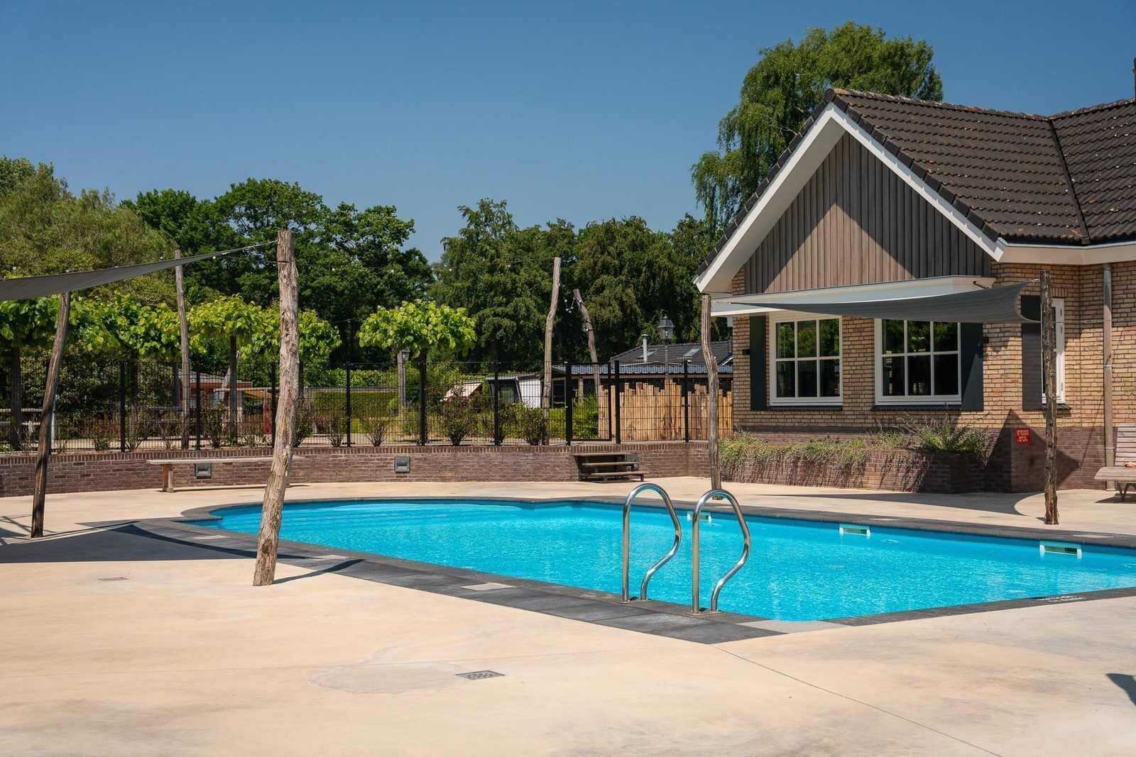 A swimming pool with metal ladders, surrounded by a paved area with trees and a building with a sloped roof.