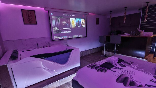 Une chambre moderne avec un grand lit, une baignoire intégrée et un téléviseur fixé au mur diffusant un film.