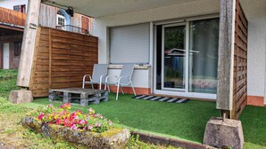 Un espace extérieur abrité doté d’une terrasse en bois, de deux chaises, d’une petite table et d’une porte-fenêtre coulissante donnant sur un jardin.