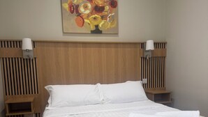 Un lit bien fait, avec du linge de lit blanc, une tête de lit en bois et un tableau de fleurs accroché au mur.