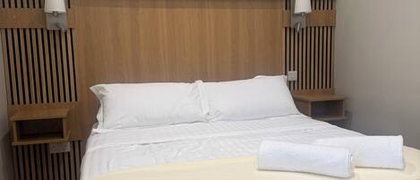 A neatly made bed with white linens, a wooden headboard, and a painting of flowers on the wall.