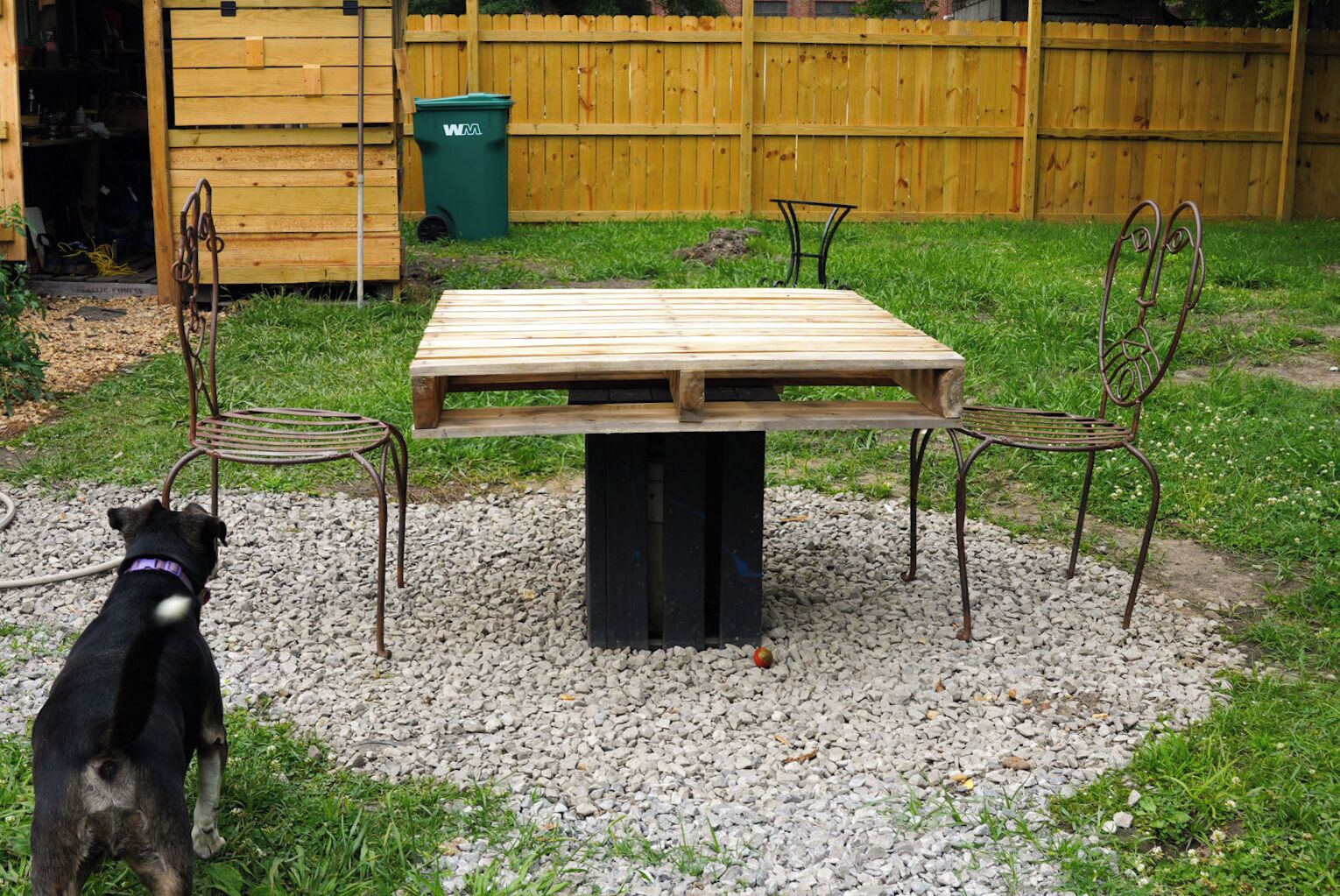A wooden picnic table with metal chairs in a backyard with a gravel ground, a wooden fence, and a green trash bin.