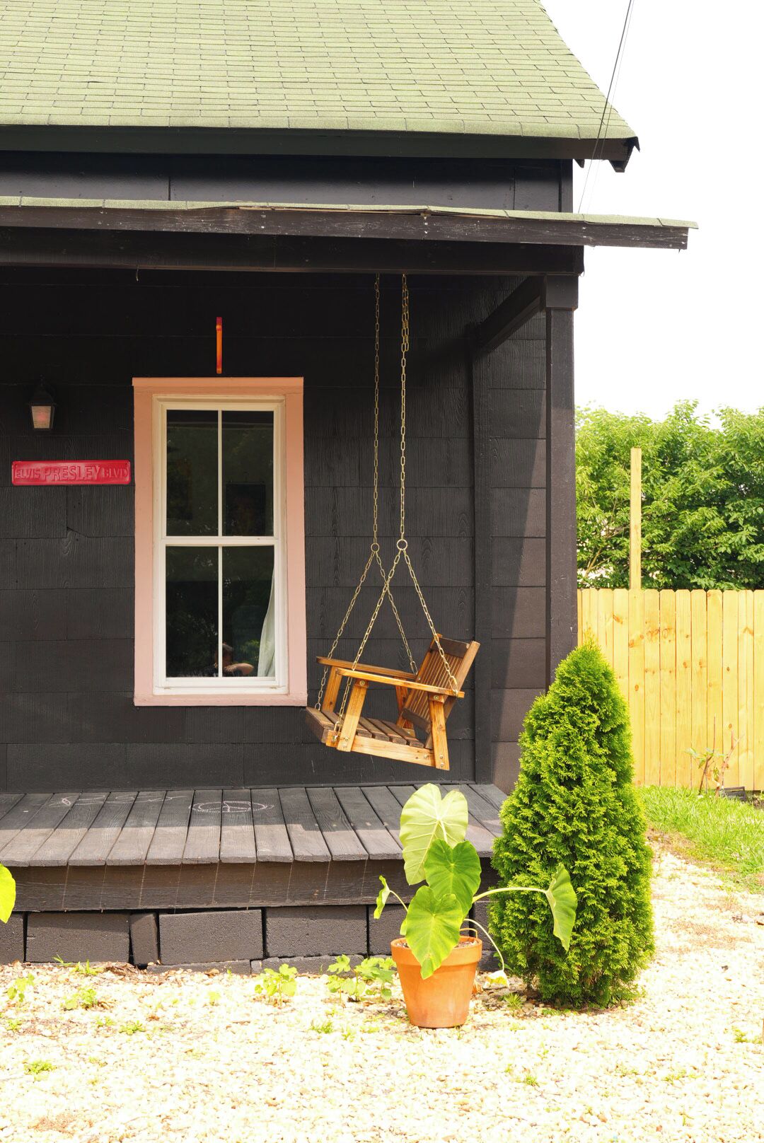 A dark wooden house with a porch, a swing chair, a potted plant, and a small tree.