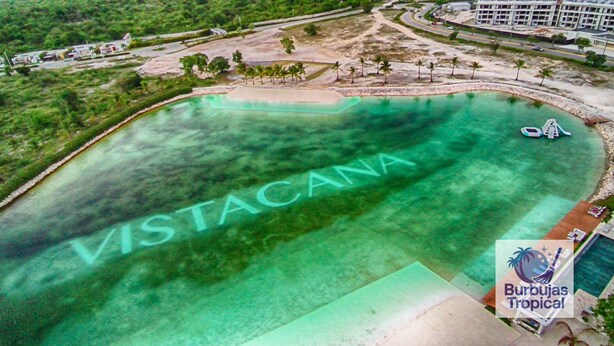 An aerial view of a beach with the word "VISTACANA" written in the water. There are palm trees and a building in the background.