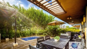 An outdoor patio area with a pool, wooden furniture, and lush greenery.