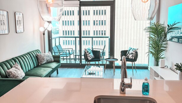 Eine moderne Küche mit Spüle, ein Essbereich mit einem grünen Sofa und Stühlen, und Blick auf eine Stadtlandschaft durch große Fenster.
