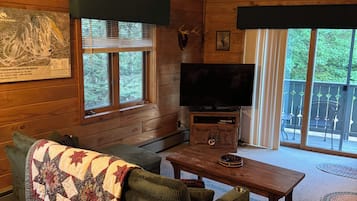 L’intérieur d’un chalet accueillant, avec un plafond en bois, un téléviseur, une cheminée et une grande fenêtre.