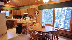 A wooden cabin kitchen with a dining area, wooden cabinets, a stove, and a large window with a view of trees.