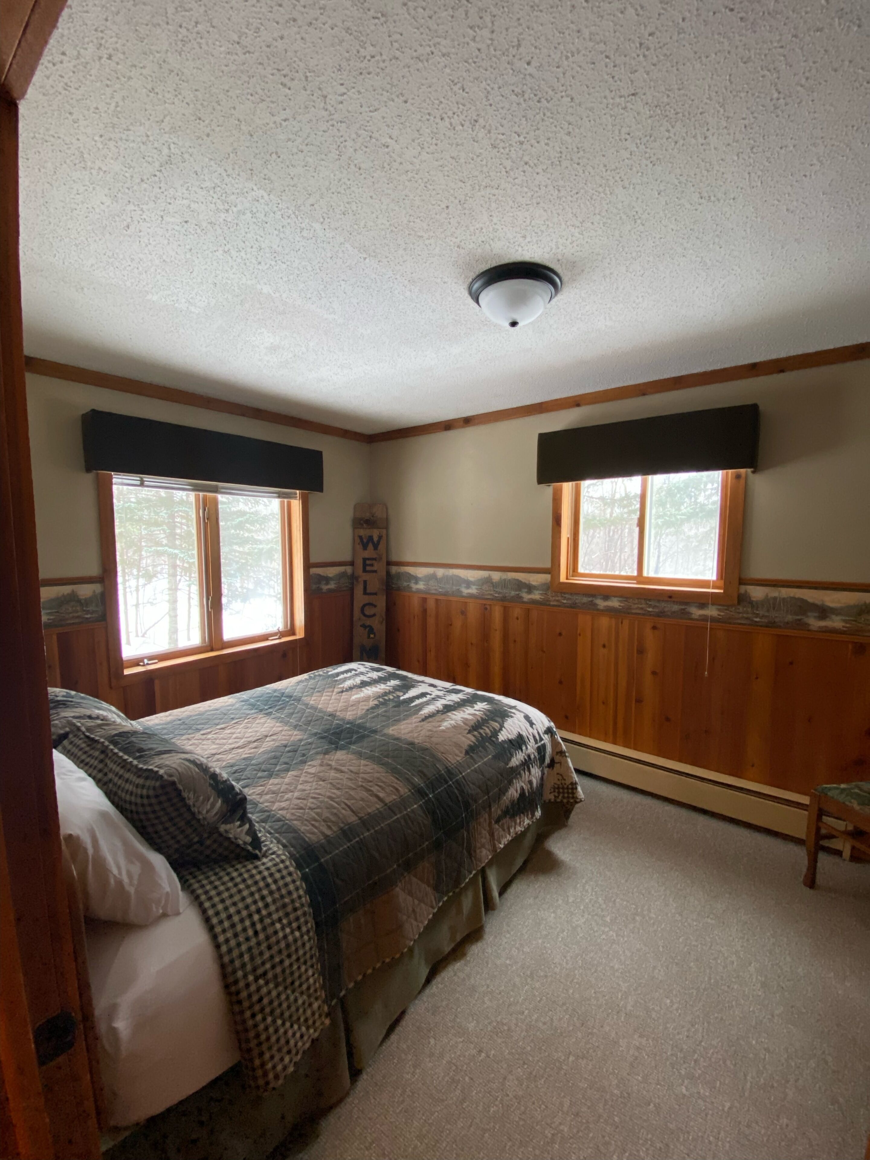 Une chambre à coucher avec un lit, deux fenêtres, des murs en bois, un luminaire au plafond et une pancarte indiquant « BIENVENUE ».