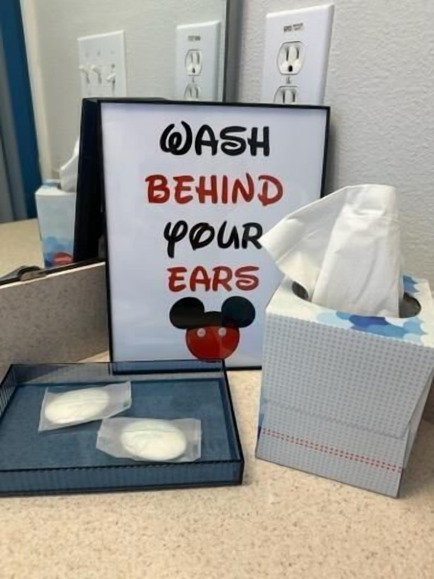 Ein Schild mit einem Mickey-Mouse-Emoji und dem Text 'HINTER DEN OHREN WASCHEN' neben einer Taschentuchbox und einem Spender.
