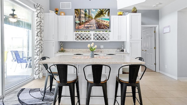A modern kitchen with a central island, white cabinetry, and a large window overlooking a beach scene. - Emerald Island Resort 6 Bed Pool & Spa-10min to Disney! (Kissimmee)
