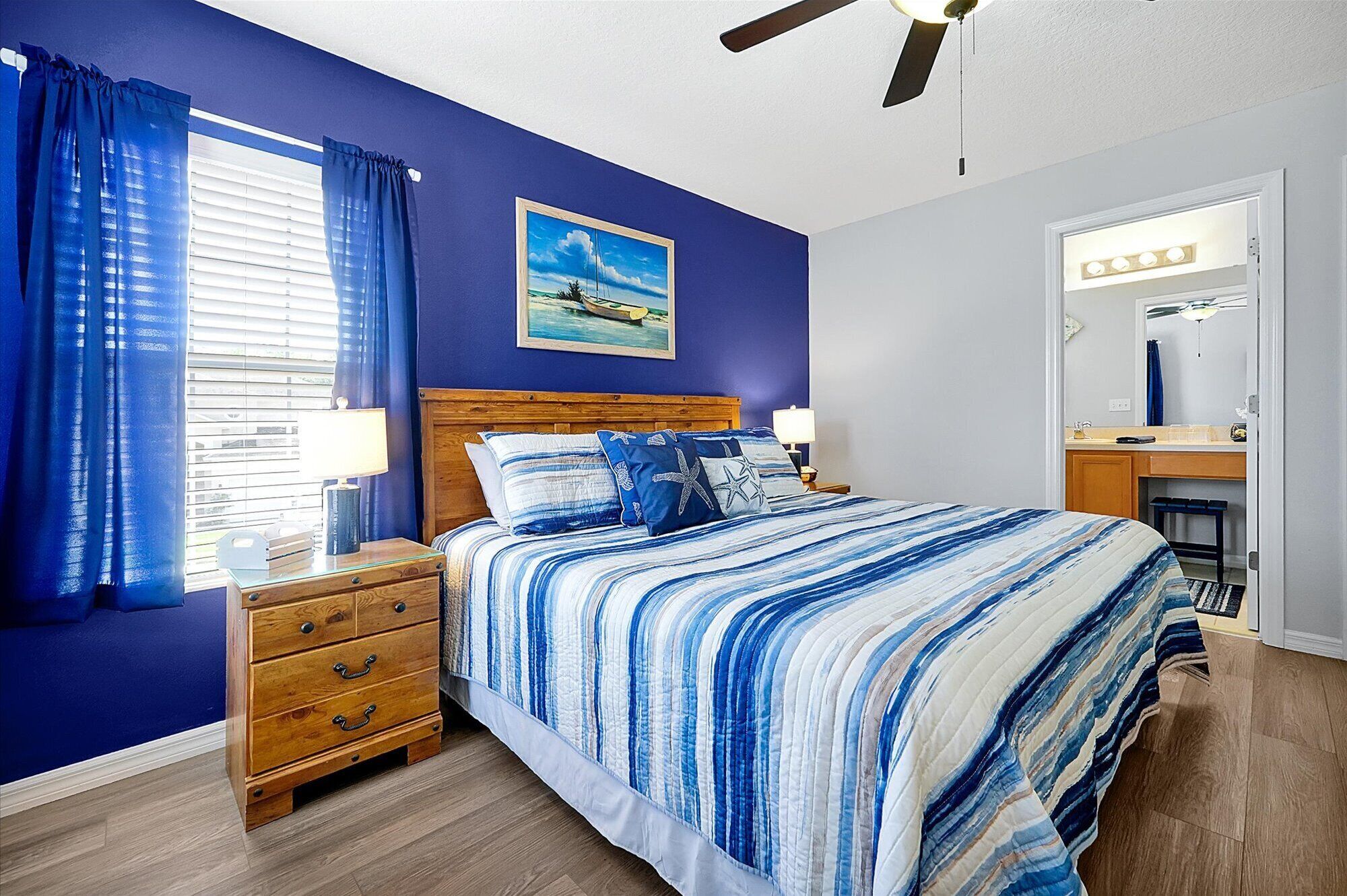 Une chambre à coucher avec un lit en bois, une table de chevet, une commode, une fenêtre avec des rideaux, un ventilateur de plafond, un tableau et une salle de bain avec un miroir et des luminaires.
