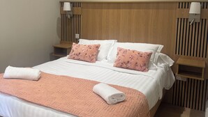 A neatly made bed with a peach-colored blanket and pillows, a wooden headboard, and a painting of flowers on the wall.