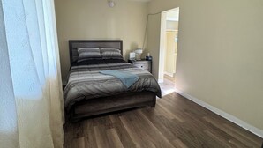 Un dormitorio con piso de madera, una cama con cubrecama a rayas, una mesita de noche con lámpara y una ventana con cortinas traslúcidas.