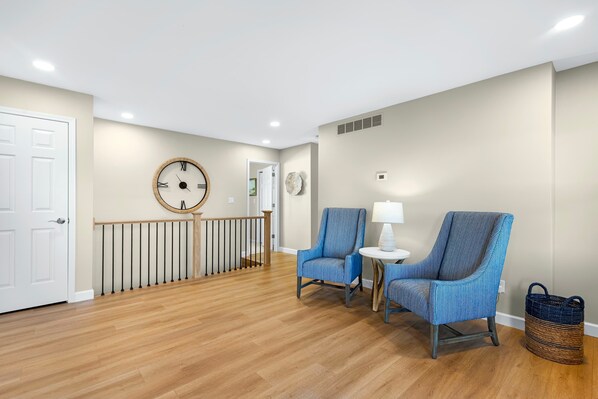 A spacious room with a wooden floor, a white door, a blue armchair, a small table with a lamp, and a wicker basket. - Panoramic Paradise - Lakefront Home for 15 (Rocky Mount)