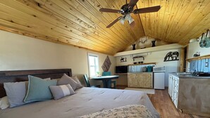 Una habitación acogedora con una cama grande, un ventilador de techo, paredes de madera, una pequeña cocina y una ventana.