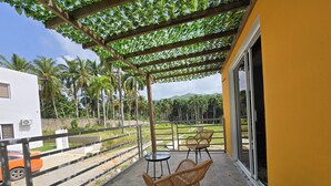 A balcony with two wicker chairs and a small table, overlooking a grassy area with palm trees and a mountain in the distance.