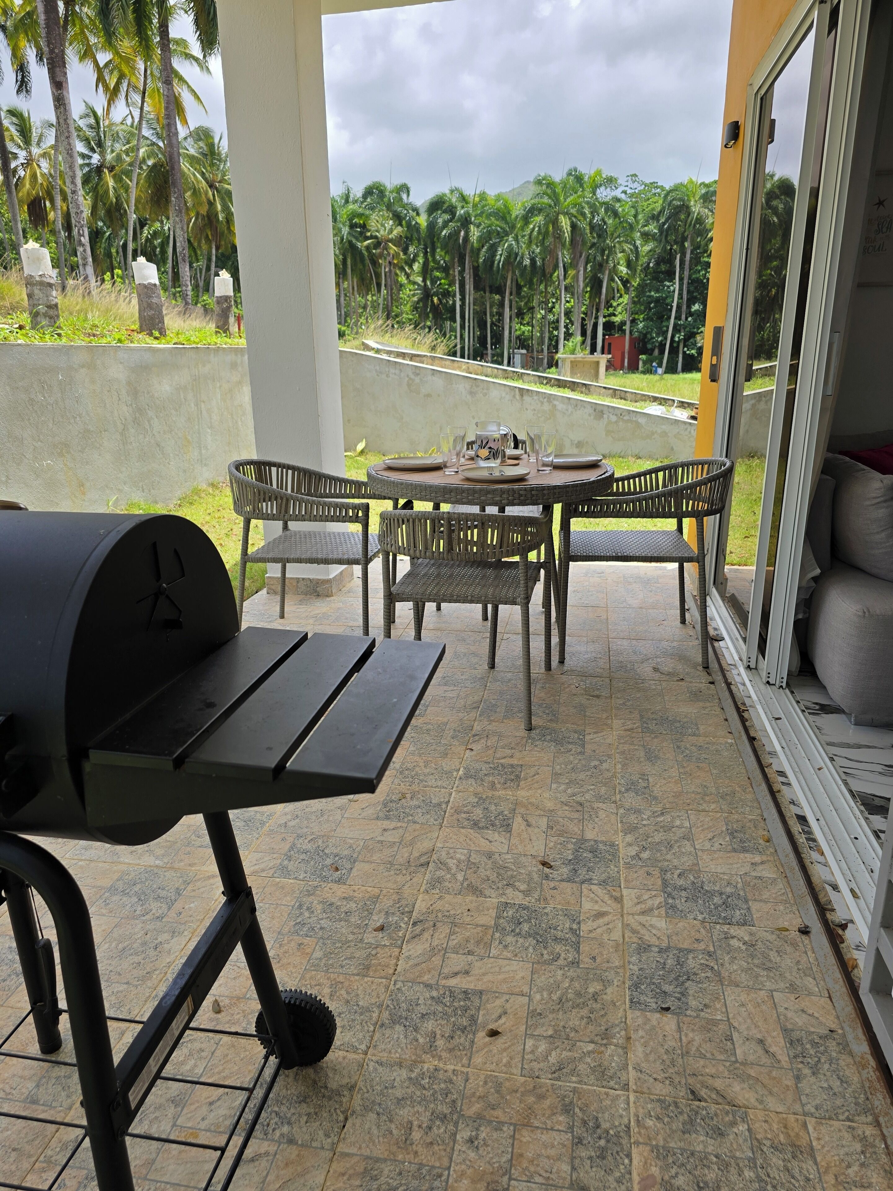 Een terras buiten met een barbecue, rieten stoelen en een eettafel.
