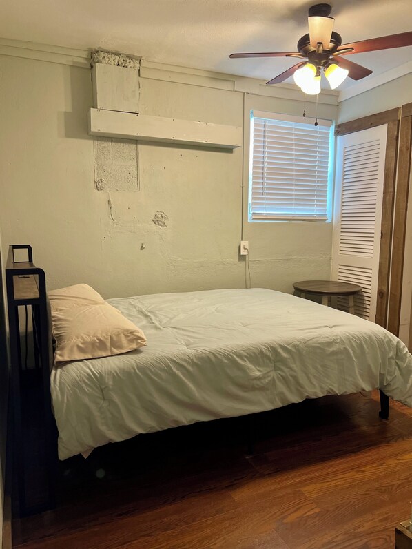 Un dormitorio con una cama individual, un ventilador de techo, una ventana con persianas y una pequeña mesa redonda.