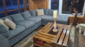 A modern living room with a grey sectional sofa, a wooden coffee table, and a large window offering a view of the outdoors. - Double room in a family home (Dalcahue)