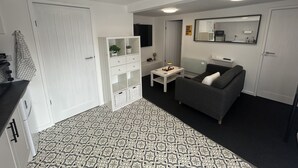 Ett kompakt bostadsutrymme med ett litet pentry, en grå soffa, ett litet bord med en lampa och ett vitt skåp med glasdörrar.