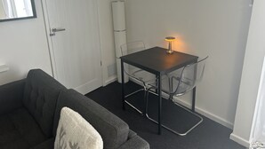 Ett litet, kompakt rum med en soffa i mörk färg, ett litet bord med en lampa och en vit dörr.