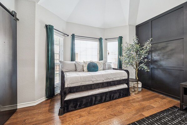 A bedroom with a large bed, a window with curtains, a plant, and a dark wall panel.