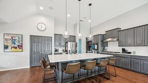 A modern kitchen with a large island, bar stools, and grey cabinetry. - Luxury 4BR Smart Home w/ Pool, Spa, Media Room & Pet-Friendly Yard (Frisco)