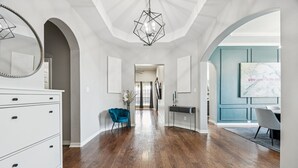 A spacious hallway with hardwood floors, white walls, and a modern chandelier. There is a blue chair, a desk, and a dining area visible. - Luxury 4BR Smart Home w/ Pool, Spa, Media Room & Pet-Friendly Yard (Frisco)