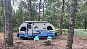 A recreational vehicle (RV) parked in a wooded area with two chairs and a table.