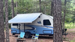 A recreational vehicle parked in a wooded area with a covered awning, two folding chairs, and a portable grill.