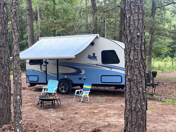 Een recreatiewagen geparkeerd in een bebost gebied, met een uitklapbare luifel, twee opklapbare stoelen en een draagbare barbecue.