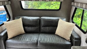 A black leather sofa with two beige pillows, a window with a view of greenery, and a patterned curtain.
