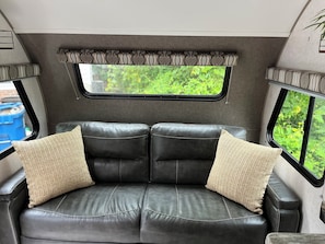 A black leather sofa with two beige pillows, a window with a view of greenery, and a patterned curtain.