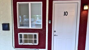 Une entrée d’immeuble avec une porte rouge portant le numéro 10, une fenêtre avec des stores et un climatiseur fixé au mur.