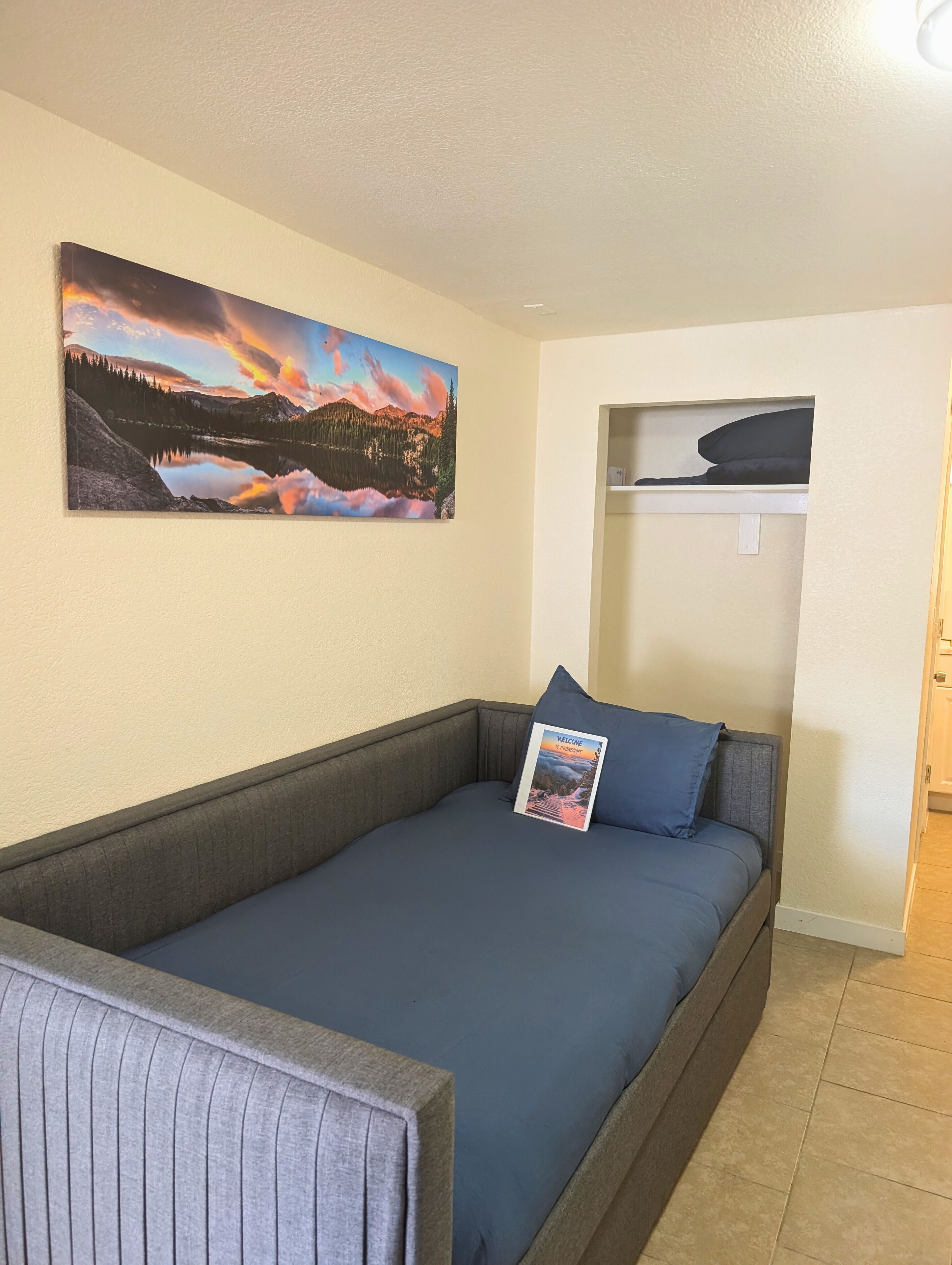 A room with a bed, a pillow, a welcome card, a painting of a landscape, and a shelf.