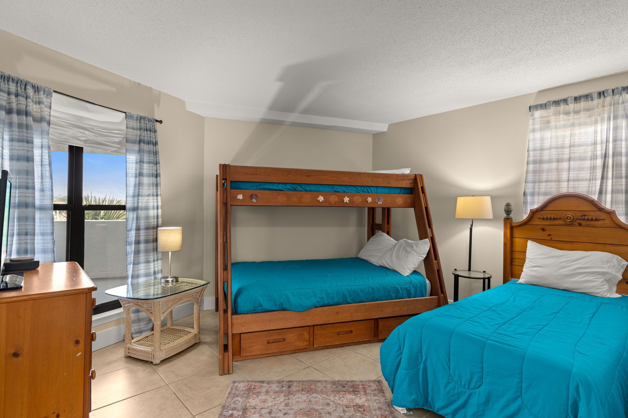 A room with a bunk bed, a desk with a lamp, a chair, and a view of the outdoors.