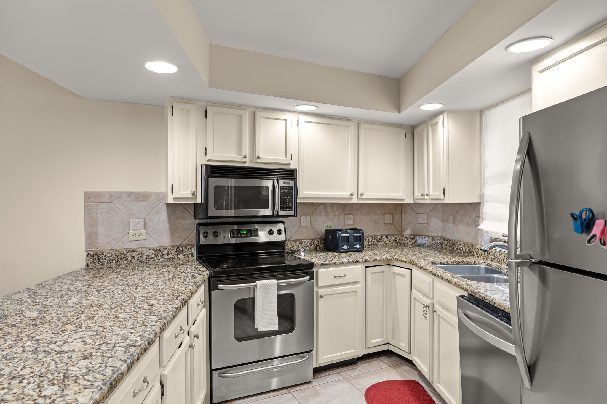 A kitchen with white cabinets, stainless steel appliances, a granite countertop, and a red rug.