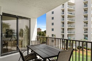 A balcony with a table and chairs, a glass door, and a view of a tall building with multiple balconies. - July 9 - 14 Now Open! Gulf View* Tram Included* Beach/Pool Access*New Bathrooms! (Miramar Beach)