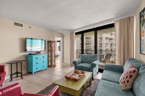 Un salon avec un téléviseur, un canapé bleu, un fauteuil rouge, un meuble en bois et un balcon donnant sur des immeubles.