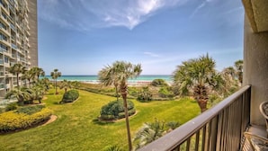 Un balcon offrant une vue sur un jardin bien entretenu, des palmiers et une plage lointaine.