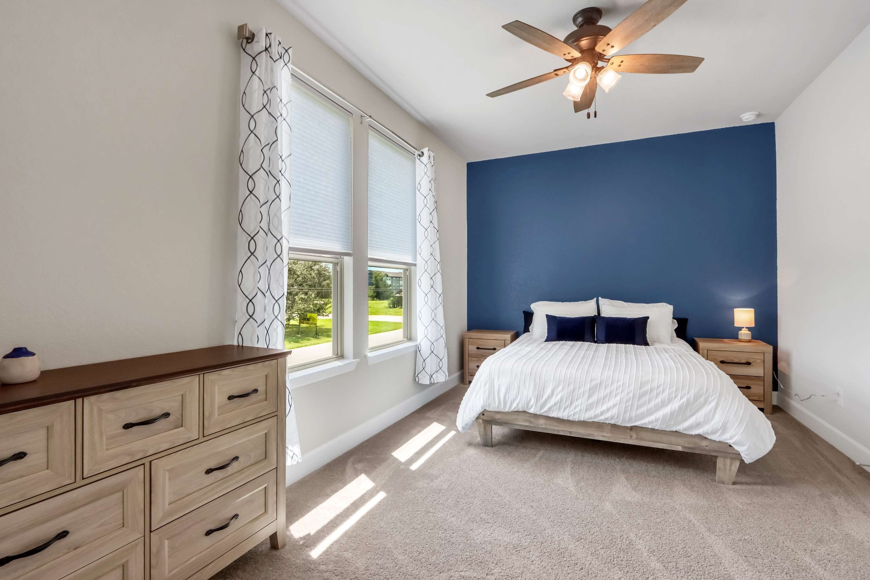 A bedroom with a bed, nightstands, a dresser, a ceiling fan, and large windows with curtains.
