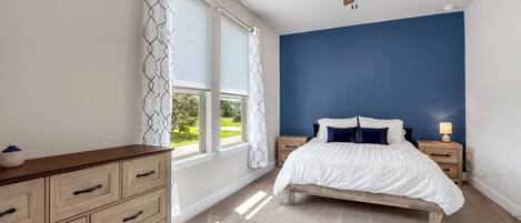 A bedroom with a bed, nightstands, a dresser, a ceiling fan, and large windows with curtains.