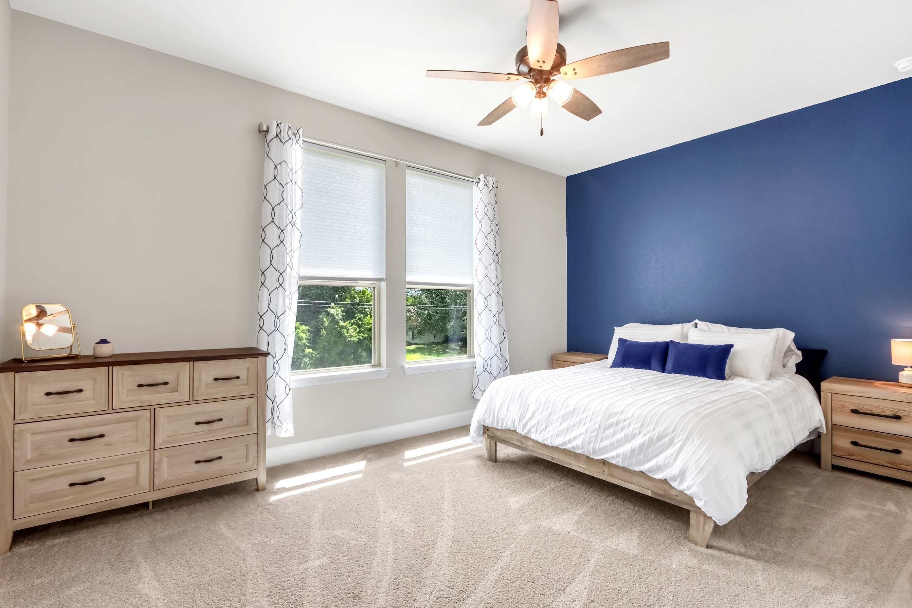 A bedroom with a bed, nightstands, a dresser, a ceiling fan, and large windows with curtains.